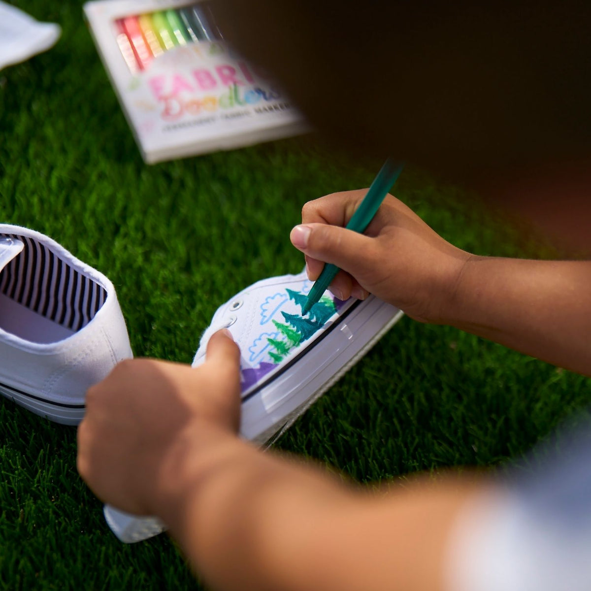 Person painting a white shoe with green paint on grass, with fabric paint book in the background.