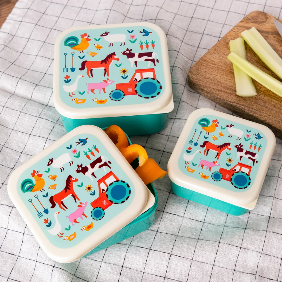 Three colorful children&#39;s lunch boxes with farm animal designs on a checkered tablecloth.