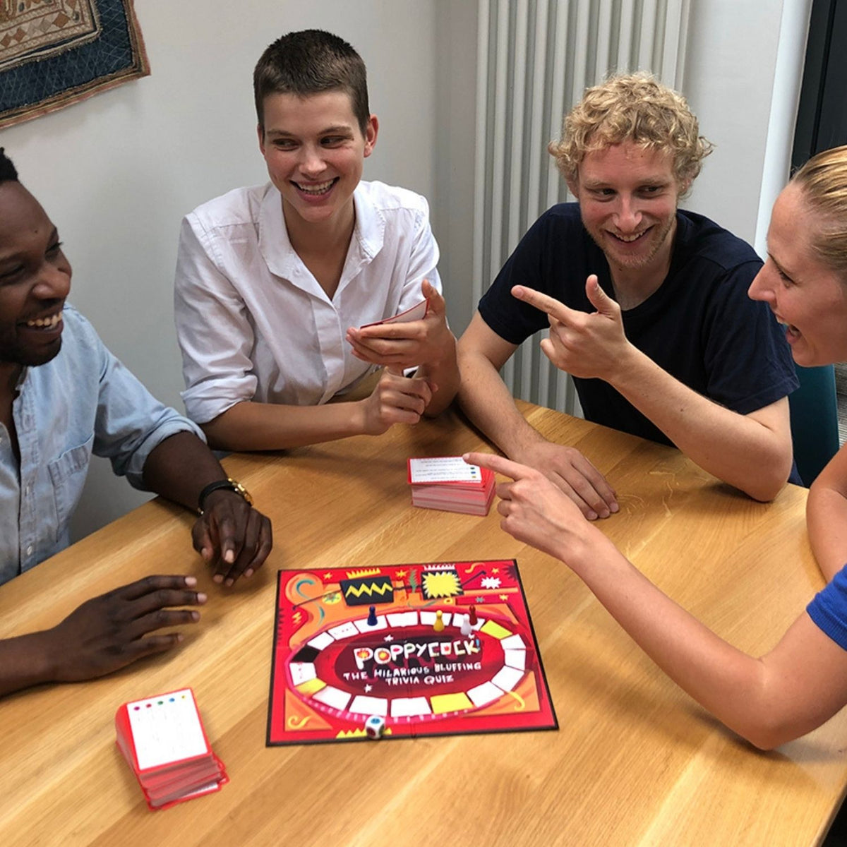 Four people playing a board game around a table with a colorful game box on the table.