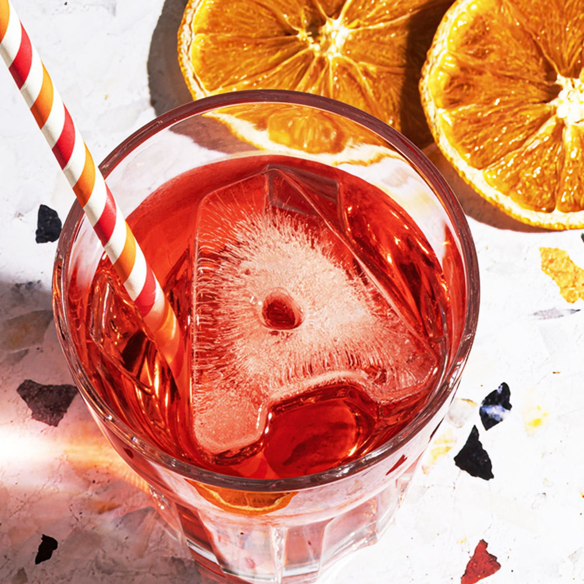 Red cocktail with a large ice cube in a glass, surrounded by orange slices on a light background.