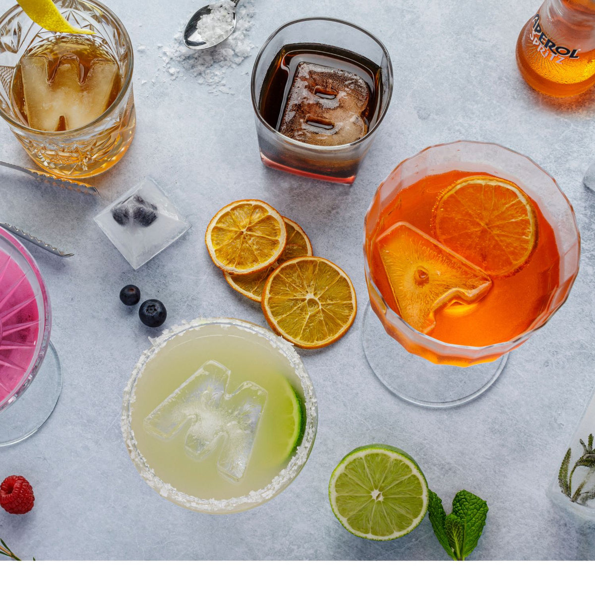 Assorted cocktails with ice cubes, fruit slices, and garnishes on a light gray surface.