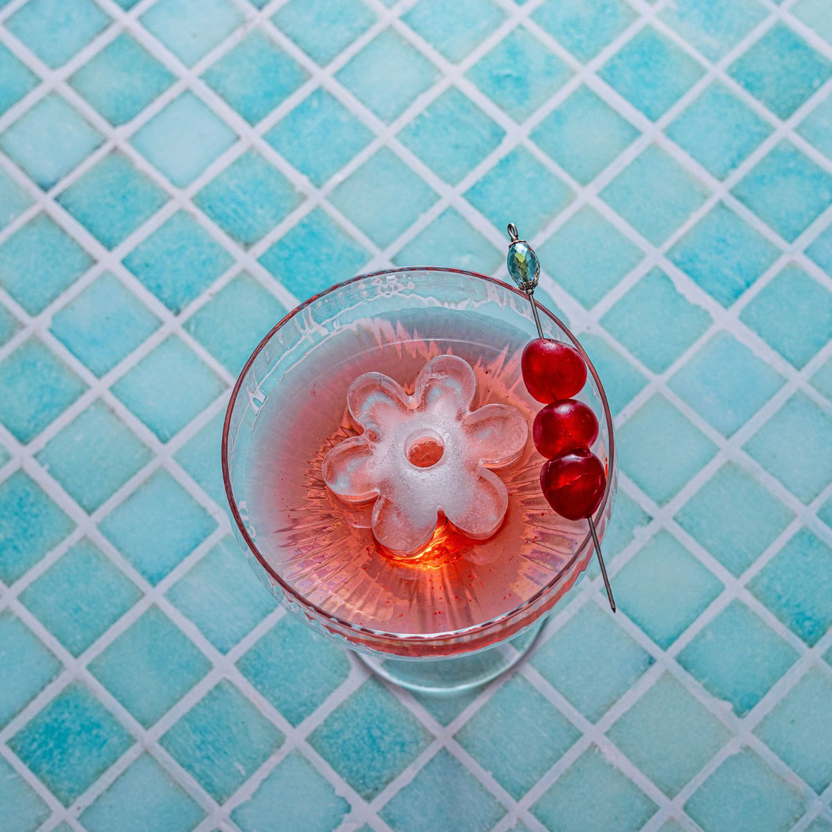Cocktail with a floral ice cube and cherry on a blue tiled surface