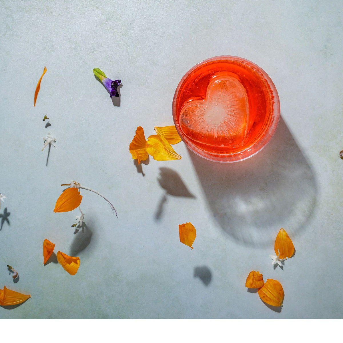 Cocktail with a heart-shaped ice cube in a red glass, surrounded by flowers on a light blue background