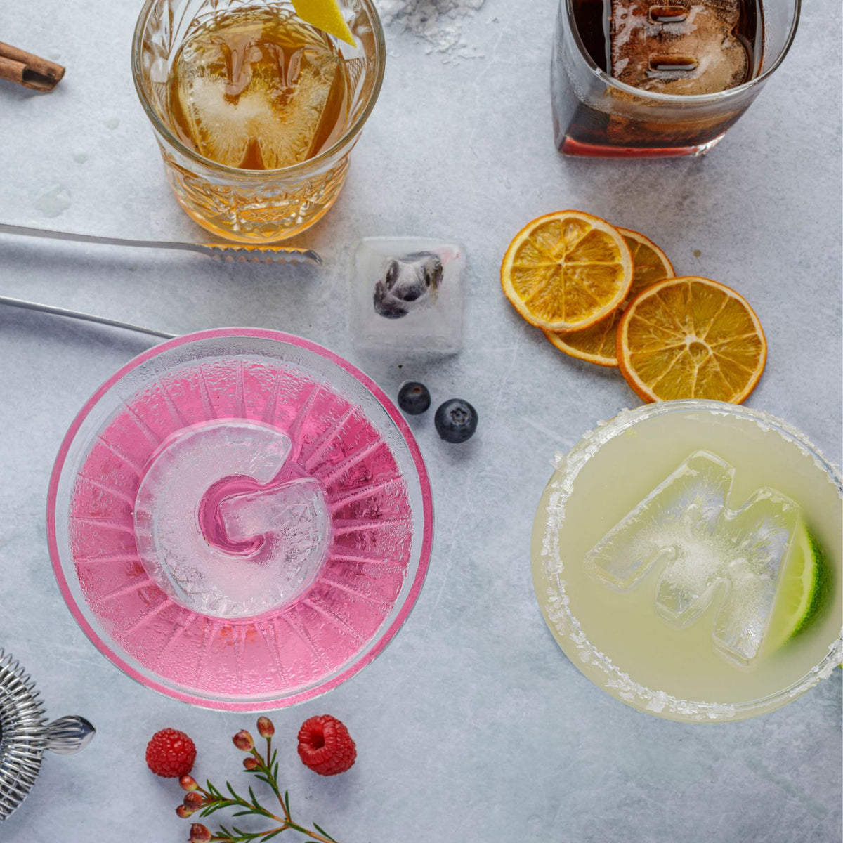 Cocktails with ice cubes, fruits, and a pink letter &#39;G&#39; ice cube tray on a light surface.