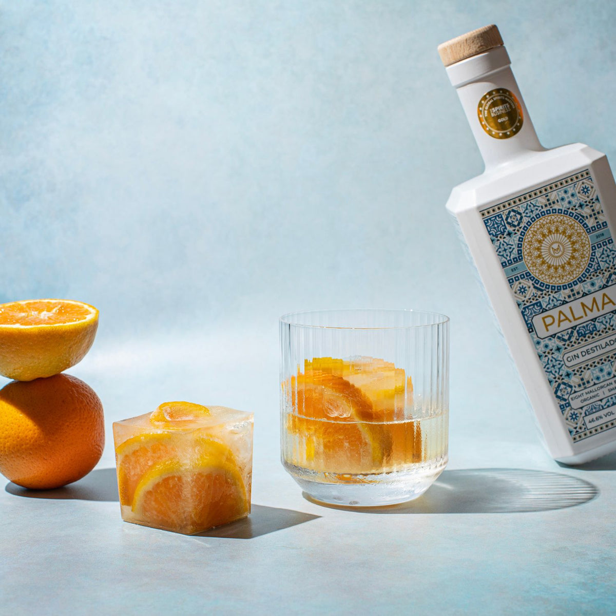 Bottle of Palma gin with two glasses of gin and tonic garnished with orange slices on a light blue background.