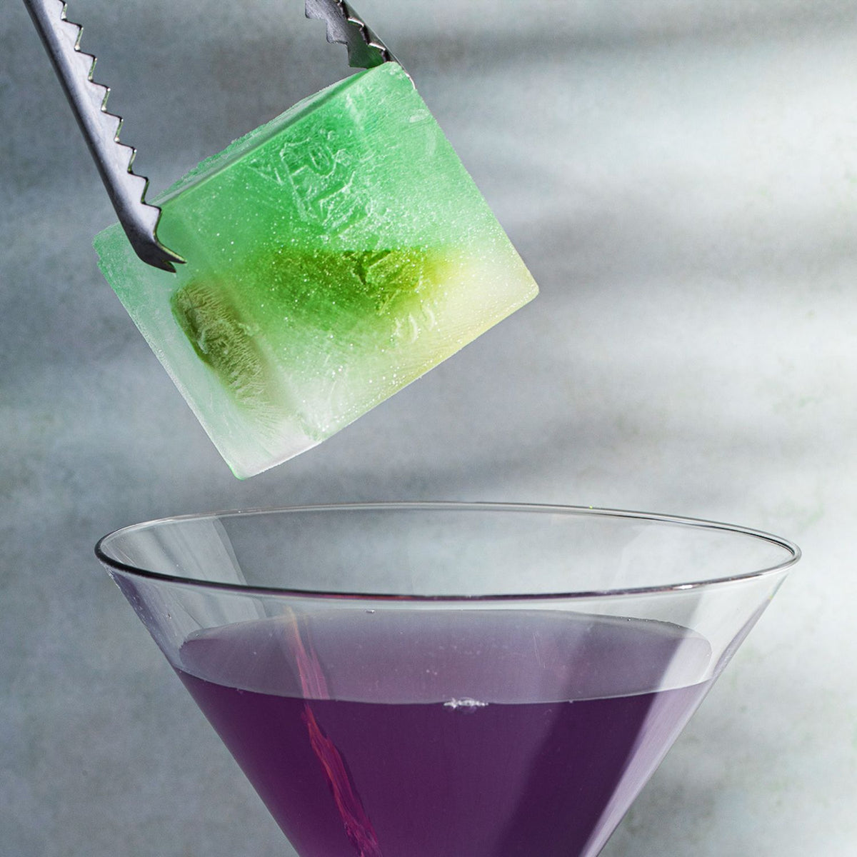 Green ice cube being removed from a purple cocktail with tongs.
