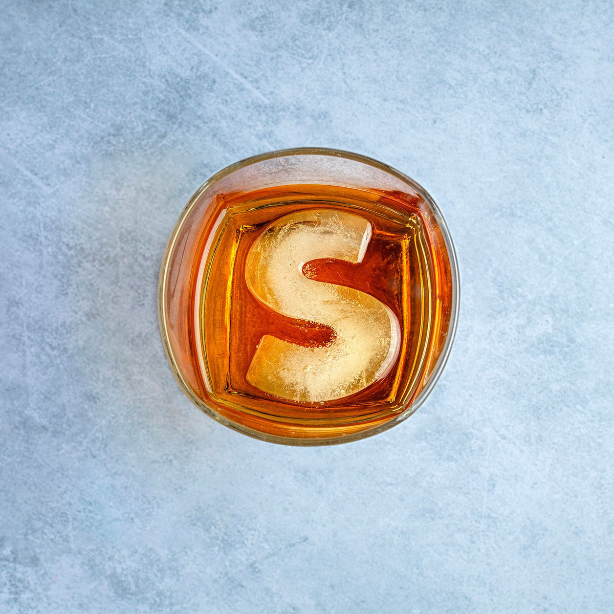 Glass of amber liquid with a large ice cube shaped like the letter &#39;S&#39; on a light blue textured background