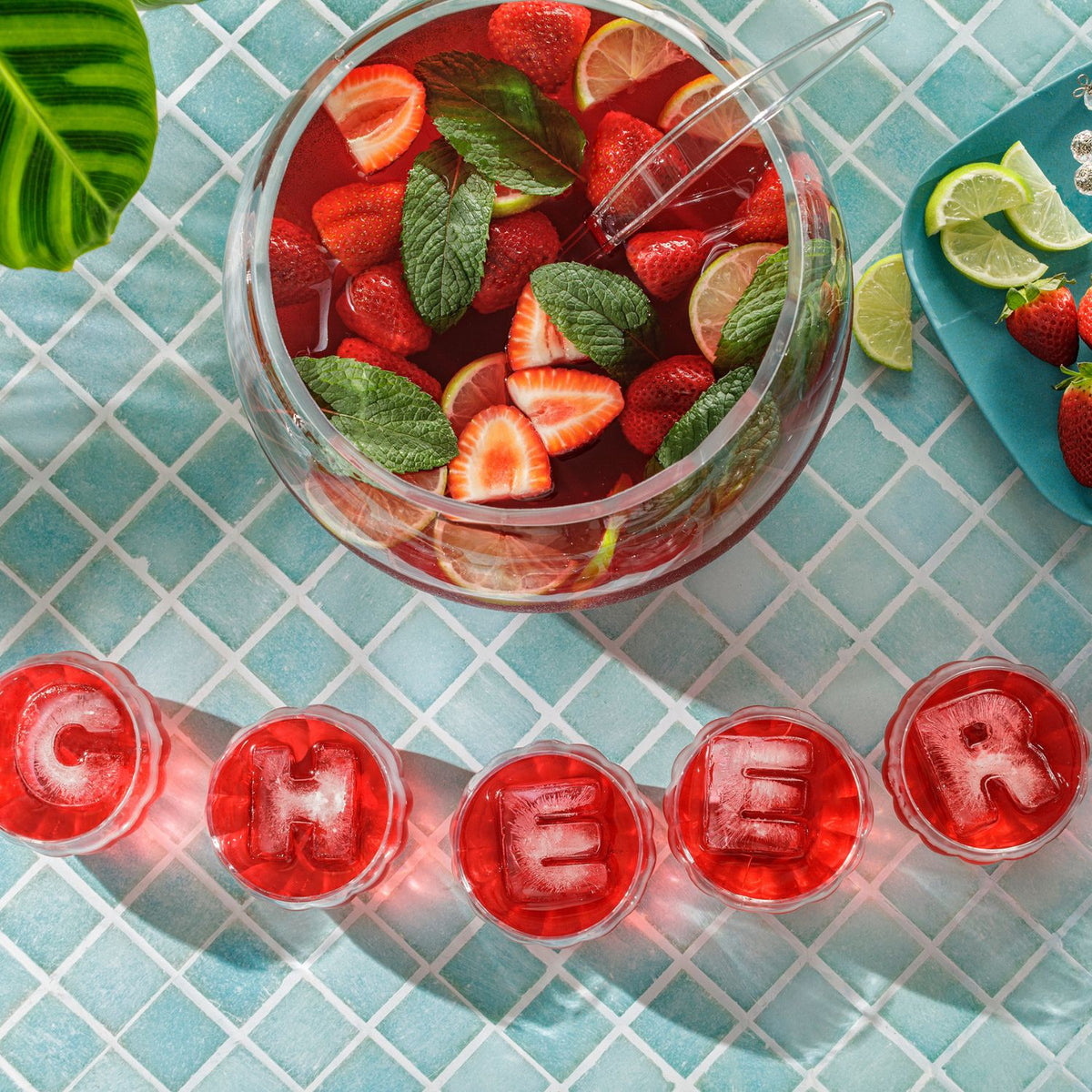 Glass punch bowl filled with strawberry lemonade on a blue tiled surface, surrounded by &#39;CHEERS&#39; letter ice cubes.