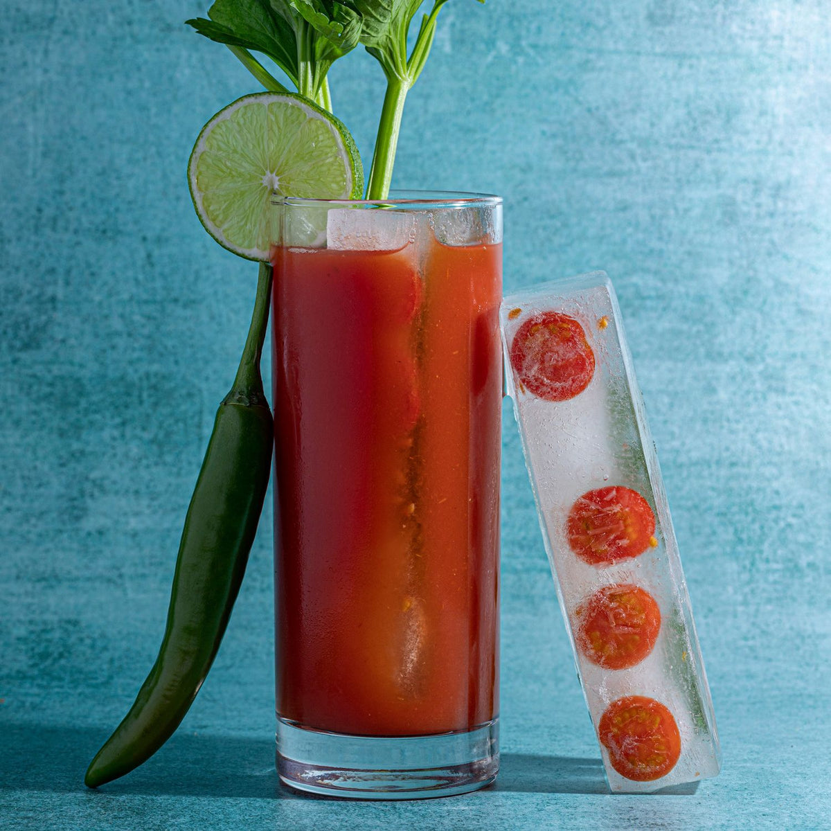 Glass of red Bloody Mary cocktail with lime, pepper, and ice cube tray on a blue background
