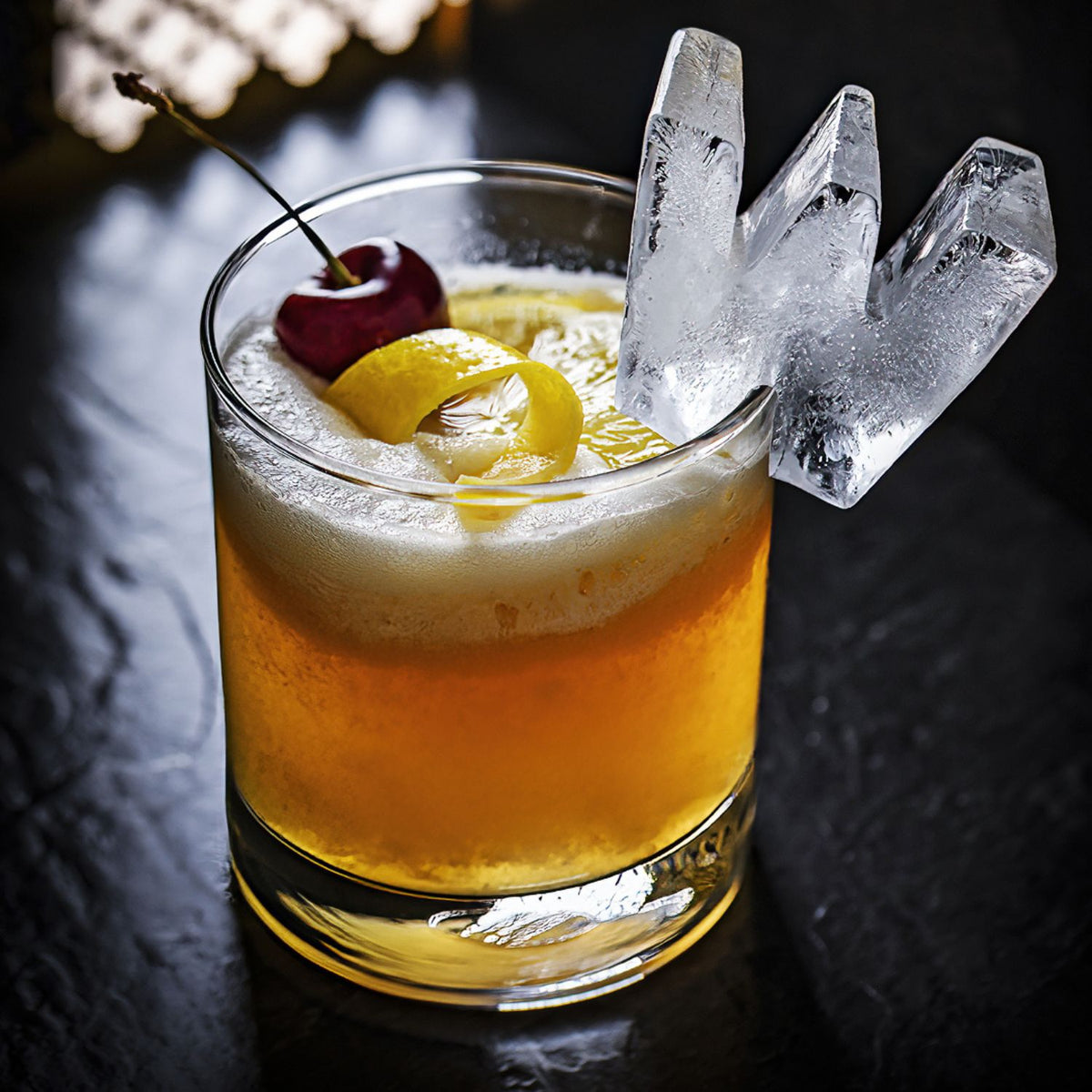 Cocktail with lemon peel, cherry, and large ice cube on a dark surface