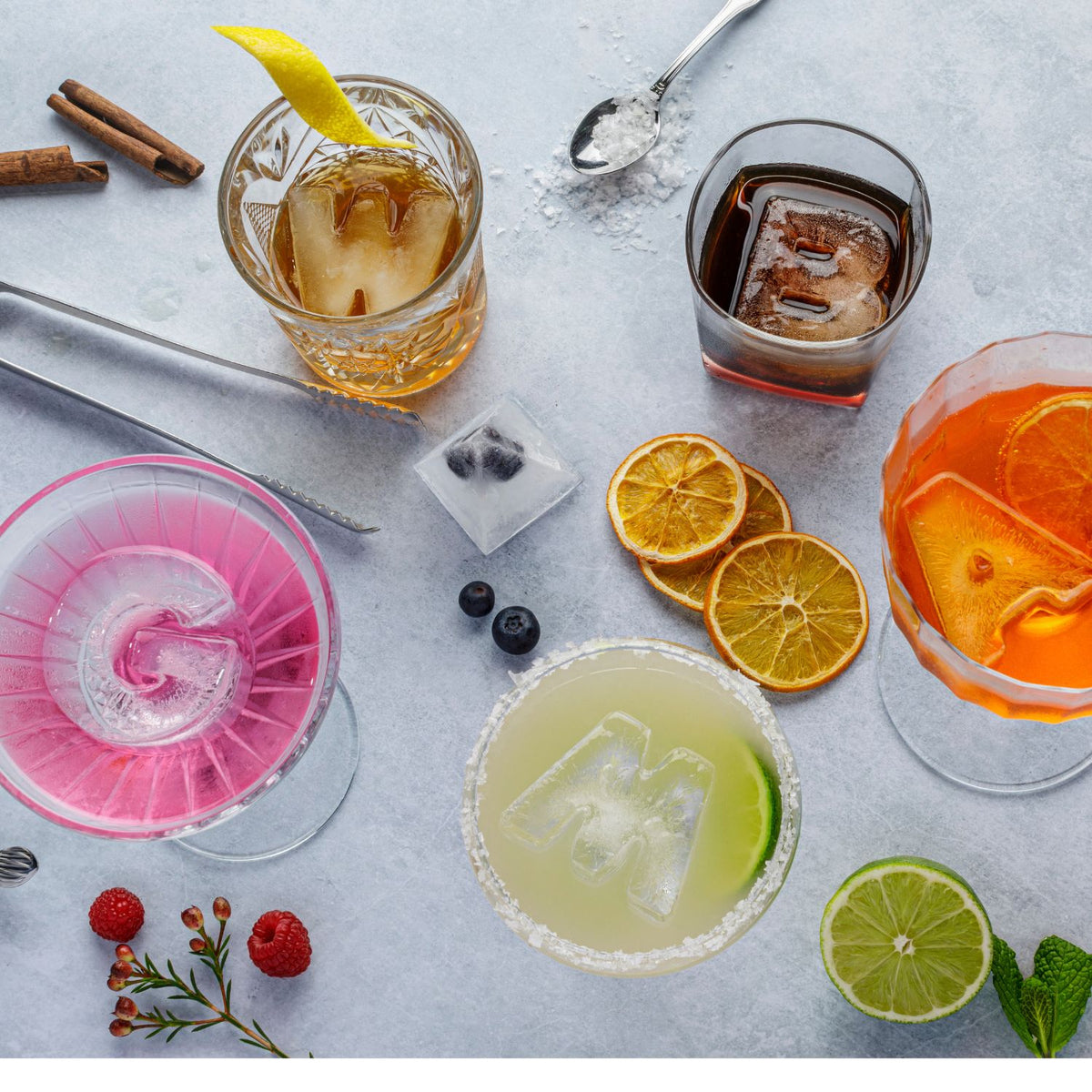 Assorted cocktails with ice cubes, fruit slices, and garnishes on a light gray surface.