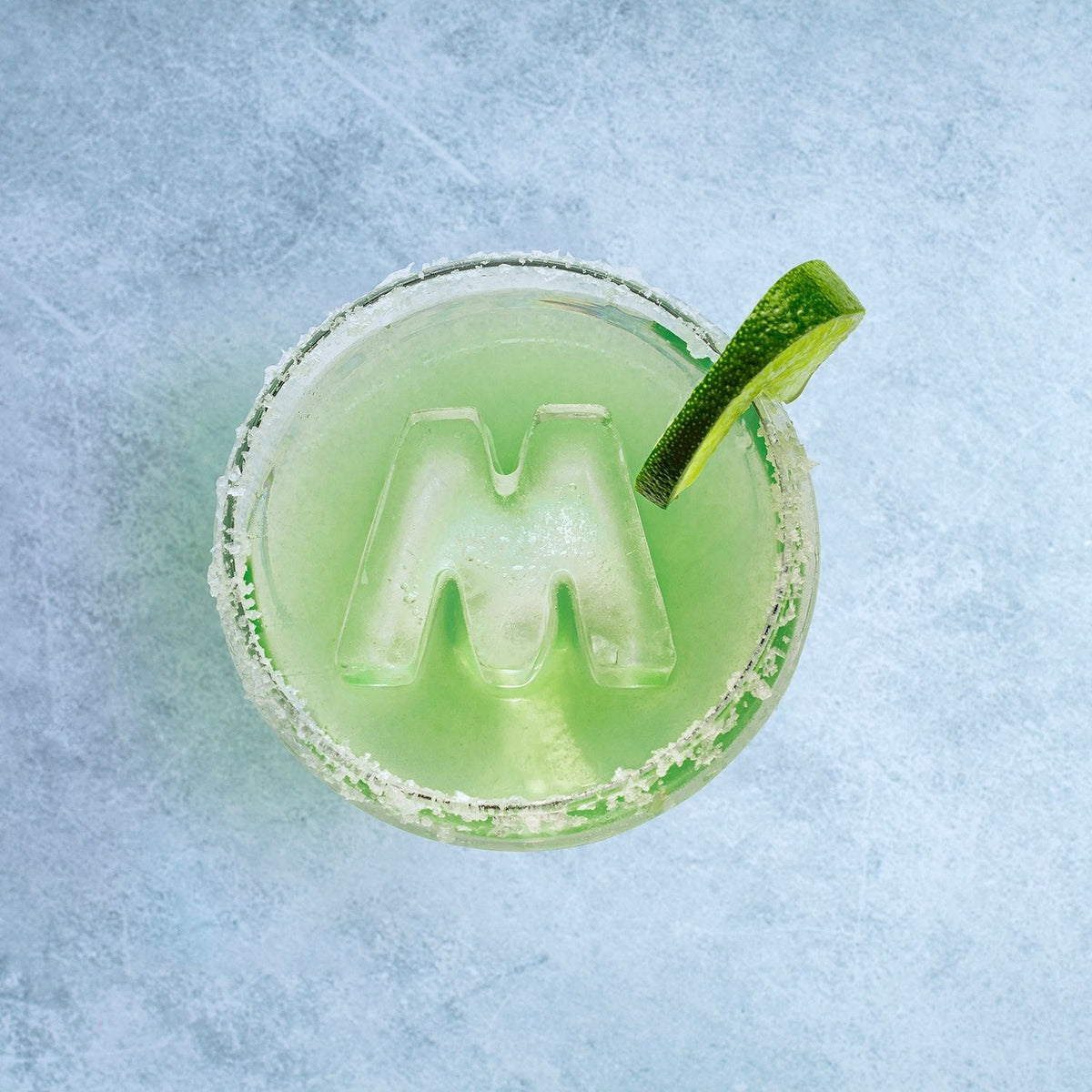 A solid, clear ice letter M being popped out of a flexible silicone M shaped ice mould, into a reen cocktail with a lime, showing the sharp, clean lines of the design. 