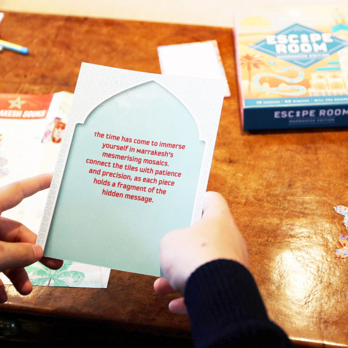 Person holding a card with text on a wooden table with an &#39;Escape Room&#39; game box in the background.