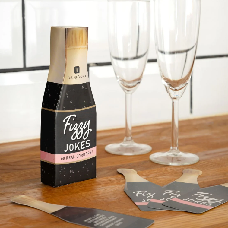 Pack of 'Fizzy Jokes' cards on a table with champagne flutes in the background