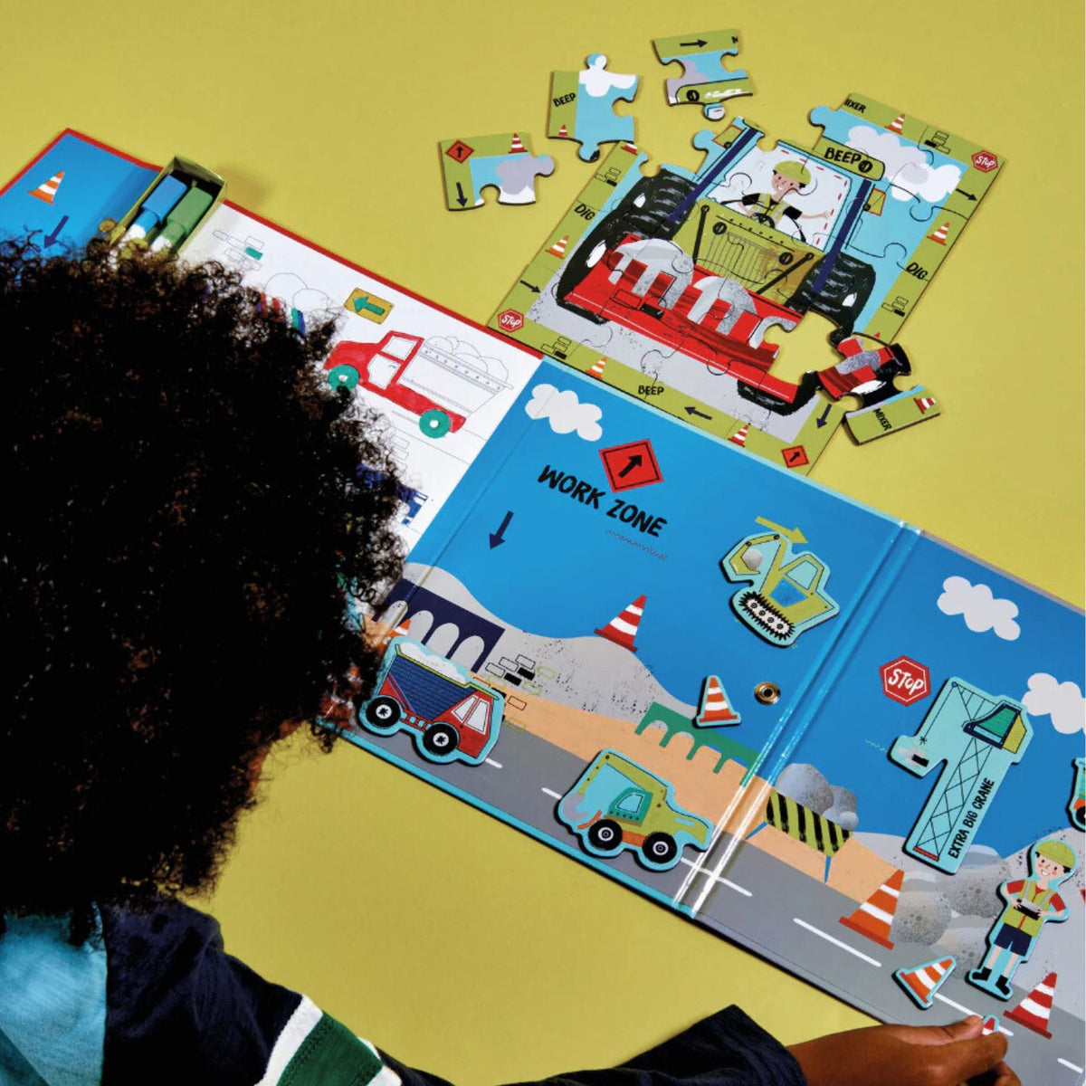 A child using a yellow dry-wipe marker on the built-in magnetic doodle board, drawing a construction site around the magnetic digger and traffic cone pieces.