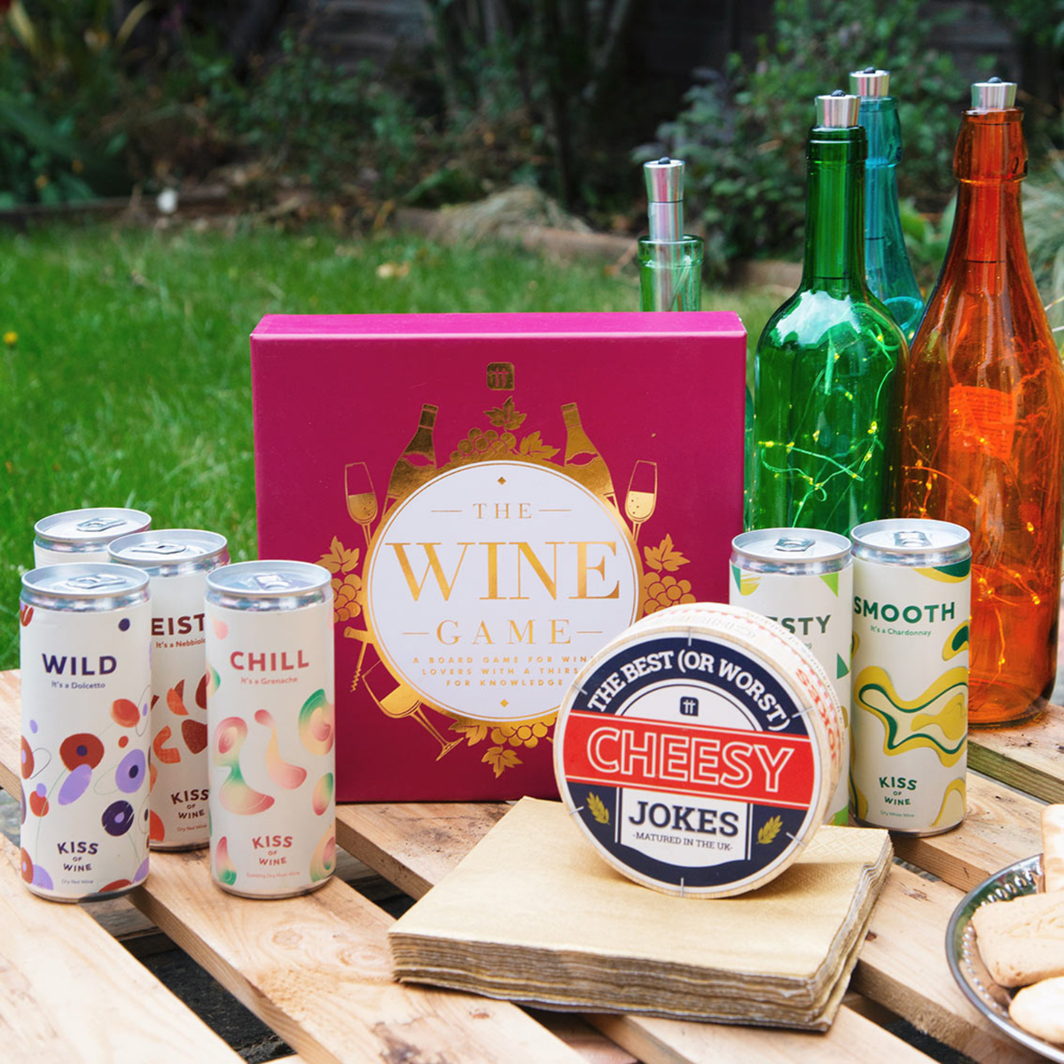 Outdoor setting with cans, 'The Wine Game' box, and decorative bottles on a wooden table.
