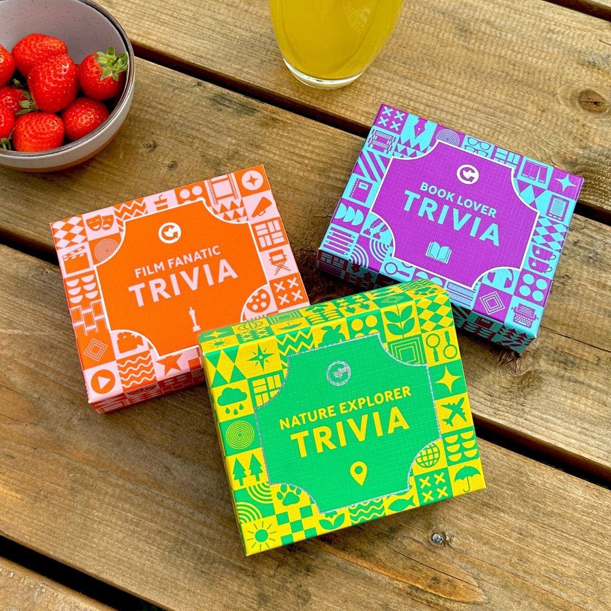 Three colorful trivia boxes, Film Fanatic Trivia, Book Lover trivia and Nature Explorer trivia on a wooden surface with strawberries and juice.
