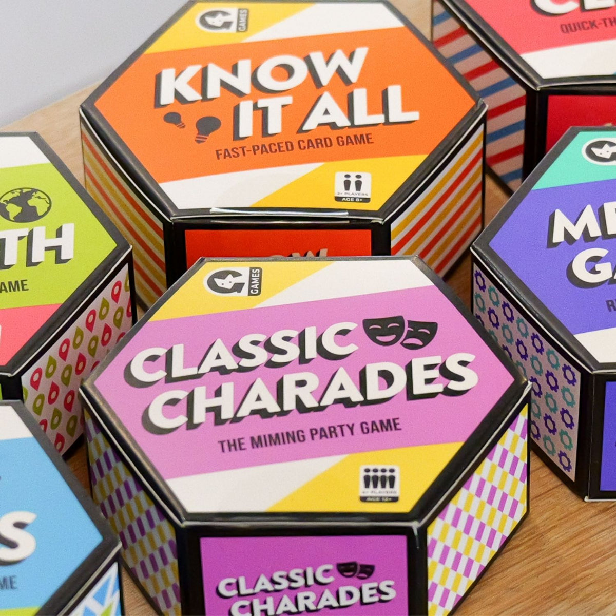 Hexagonal boxes of card games including 'Know It All' and 'Classic Charades' on a wooden surface.