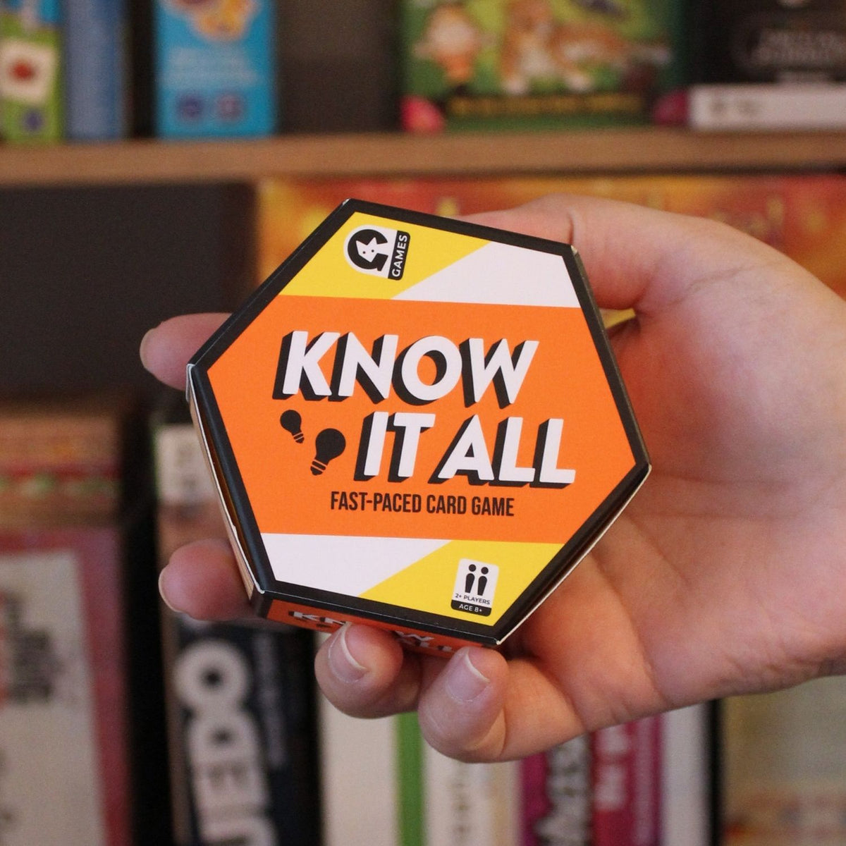 Hand holding a 'Know It All' fast-paced card game box with a bookshelf in the background