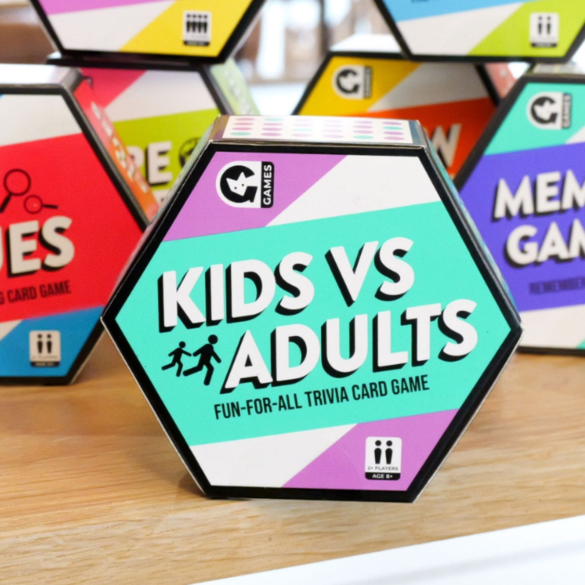 Hexagonal card game box titled 'Kids vs Adults' on a wooden surface with other colorful card game boxes in the background.