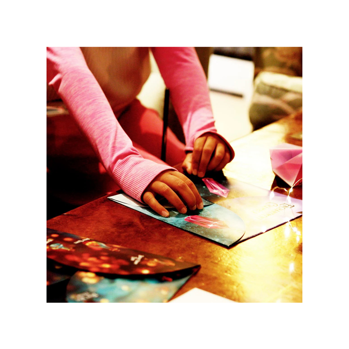 Person in pink shirt working on a craft project with colorful materials on a wooden table.