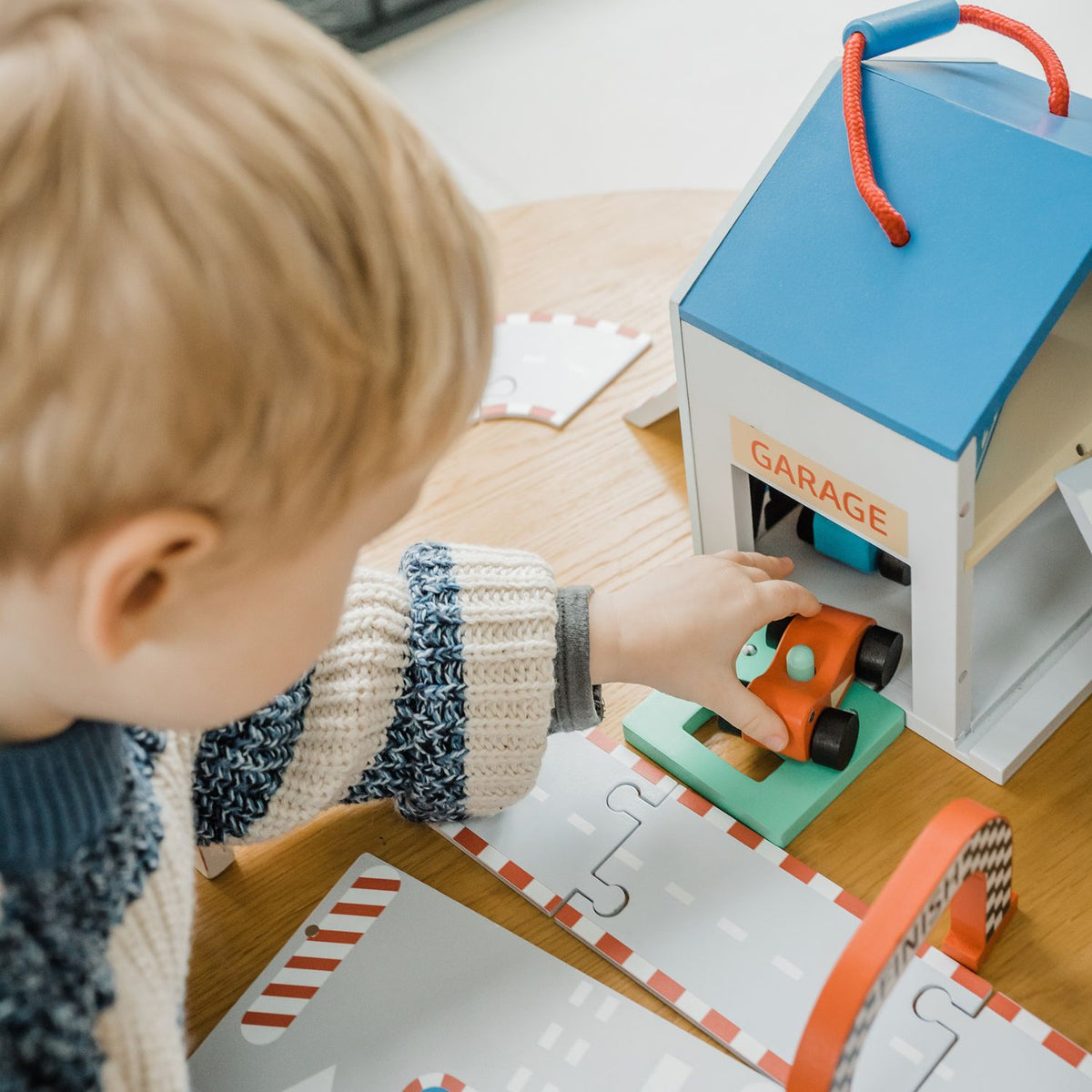 A close-up shot of a toddler’s hand zooming a red wooden race car out of the garage on the illustrated fold-out track, capturing active fine motor play.