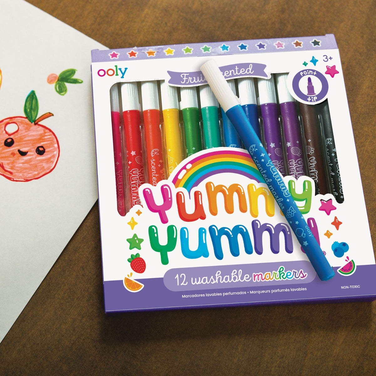 Ooly Yummy Yumm washable markers set on a wooden surface with a child's drawing of an apple.