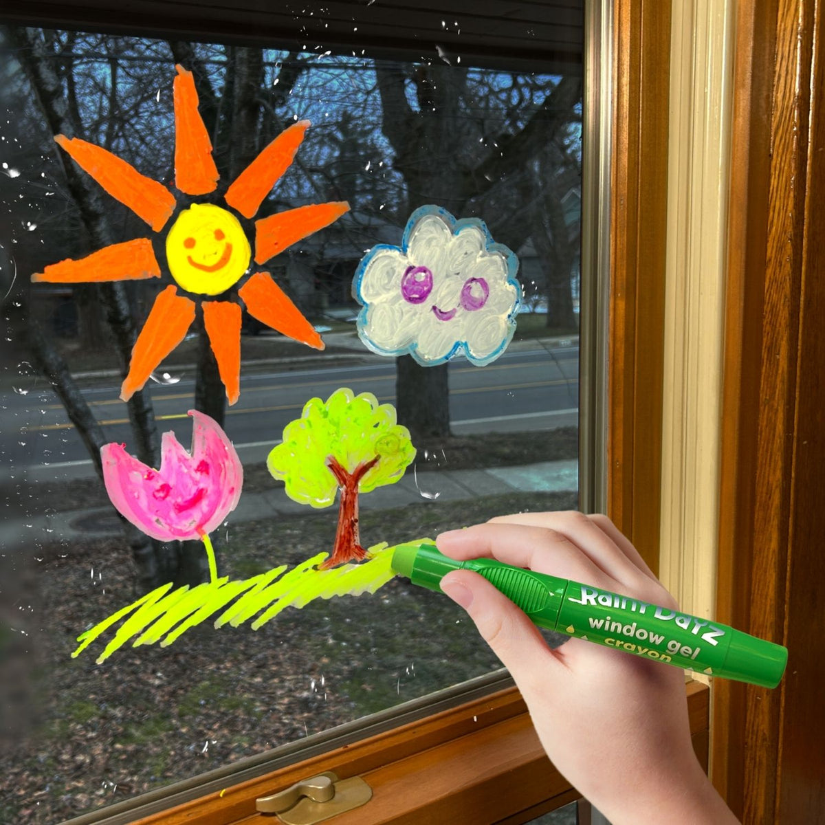 Hand holding a green window gel crayon with colorful window decorations on a glass door.