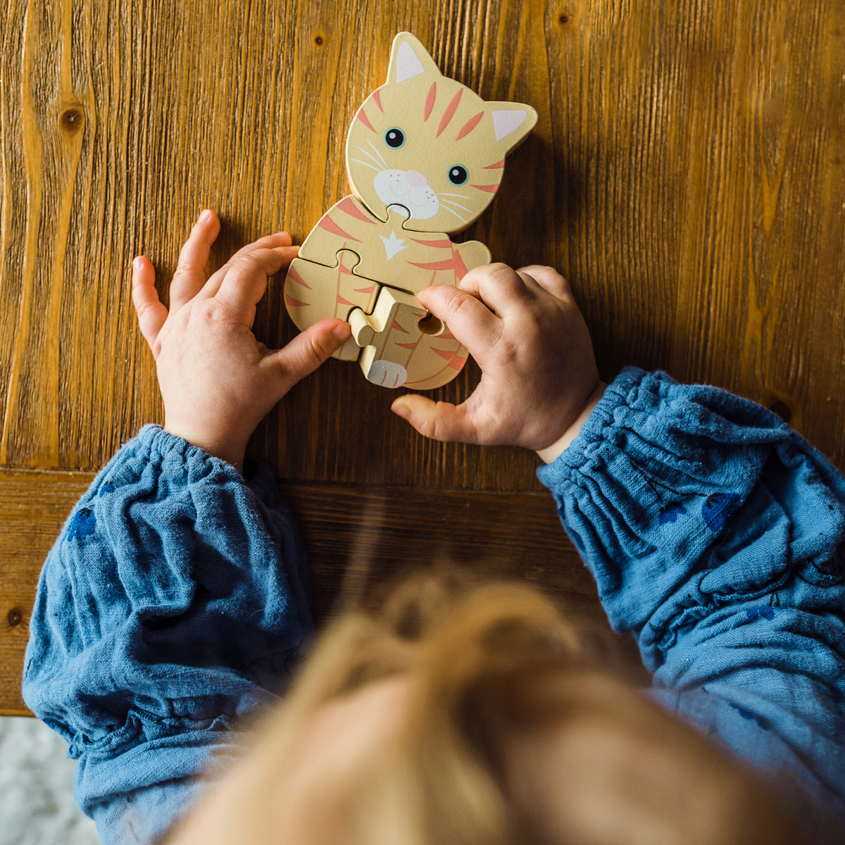 A toddler’s small hands carefully connecting two chunky wooden pieces to complete the cat puzzle on a wooden play table, illustrating fine motor dexterity.