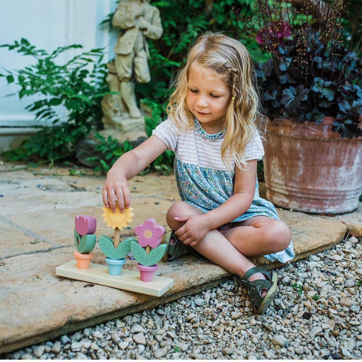 A toddler happily playing on a patio, looking proudly at the completed 3-tier wooden flower pot stacking toy.