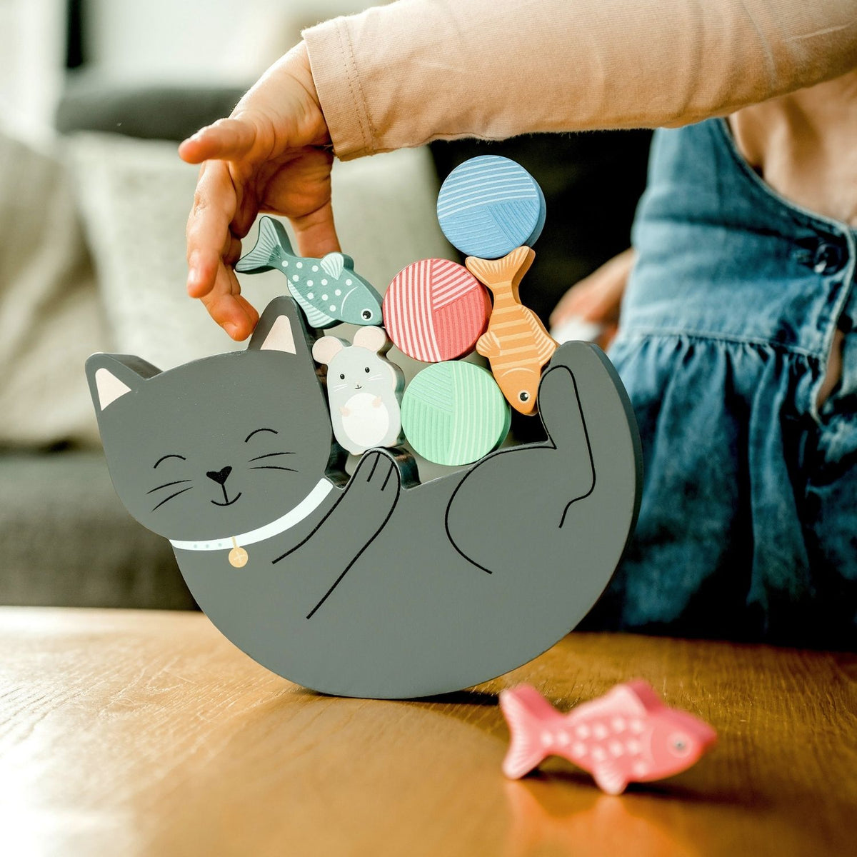 A close-up shot of a toddler’s hands carefully placing a small wooden fish on top of the precarious stacking pile, capturing focus and fine motor precision.